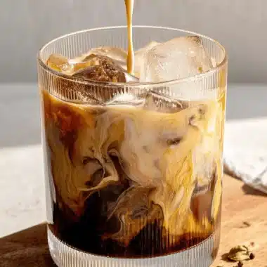 Amazing Homemade Iced Pumpkin Chai Tea Latte 4 A refreshing iced pumpkin chai tea latte being prepared, with thick cream pouring into a glass filled with ice and the dark tea mixture, creating beautiful swirls on a wooden surface with spices.