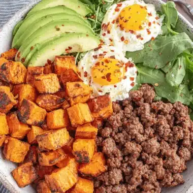 Amazing Ground Beef Bowl: Quick & Easy Dinner 3 Overhead view of a colorful ground beef bowl with sweet potatoes, fried eggs, avocado, and greens on a wooden surface.