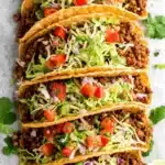 Foolproof Ground Beef Tacos for Speedy Dinner 9 Close-up of seven crispy ground beef tacos, generously filled with seasoned meat, lettuce, tomato, onion, and cheese, garnished with lime and cilantro.