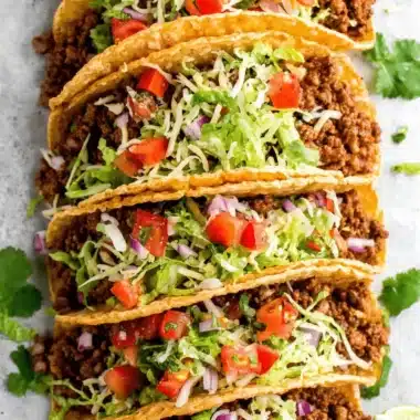 Foolproof Ground Beef Tacos for Speedy Dinner 3 Close-up of seven crispy ground beef tacos, generously filled with seasoned meat, lettuce, tomato, onion, and cheese, garnished with lime and cilantro.