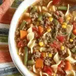 Amazing Hamburger Noodle Soup Recipe 6 Close-up overhead view of a hearty bowl of Hamburger Noodle Soup with ground beef, egg noodles, carrots, green beans, corn, and tomatoes, garnished with fresh herbs.