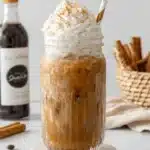 Magic Homemade Pumpkin Spice Latte Recipe 7 Iced pumpkin spice latte in a fluted glass jar, topped with whipped cream, cinnamon, and a gold-striped straw, surrounded by coffee beans and autumnal spices.