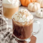 Amazing Pumpkin Spice Hot Chocolate Recipe 8 Steaming mug of pumpkin spice hot chocolate with whipped cream and cinnamon, on a wooden coaster with autumn decor.
