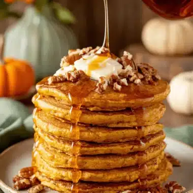 The Ultimate Fluffy Pumpkin Pancakes 3 A stack of fluffy pumpkin pancakes topped with melting butter, pecans, and maple syrup cascading down the sides, set in a warm autumnal scene.