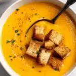Amazing Creamy Sweet Potato Soup 6 Close-up of a creamy sweet potato soup in a white bowl, garnished with croutons, parsley, and black pepper, with a ladle resting in it.