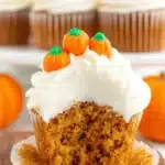 Amazing Easy Pumpkin Cupcakes Recipe 5 Close-up of a broken pumpkin cupcake revealing its moist, spiced interior, topped with creamy white frosting and candy pumpkins on a wooden board, with more pumpkin cupcakes out of focus.
