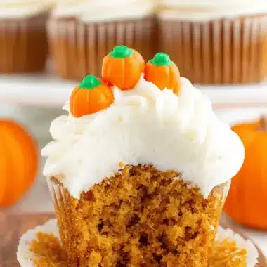 Amazing Easy Pumpkin Cupcakes Recipe 4 Close-up of a broken pumpkin cupcake revealing its moist, spiced interior, topped with creamy white frosting and candy pumpkins on a wooden board, with more pumpkin cupcakes out of focus.