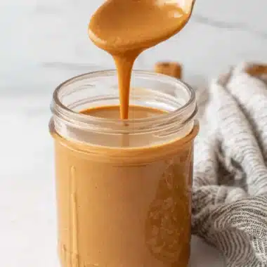 Foolproof No-Cook Pumpkin Spice Sauce Recipe 3 A silver spoon drips creamy pumpkin spice sauce into a glass jar, highlighting its smooth texture on a light background.